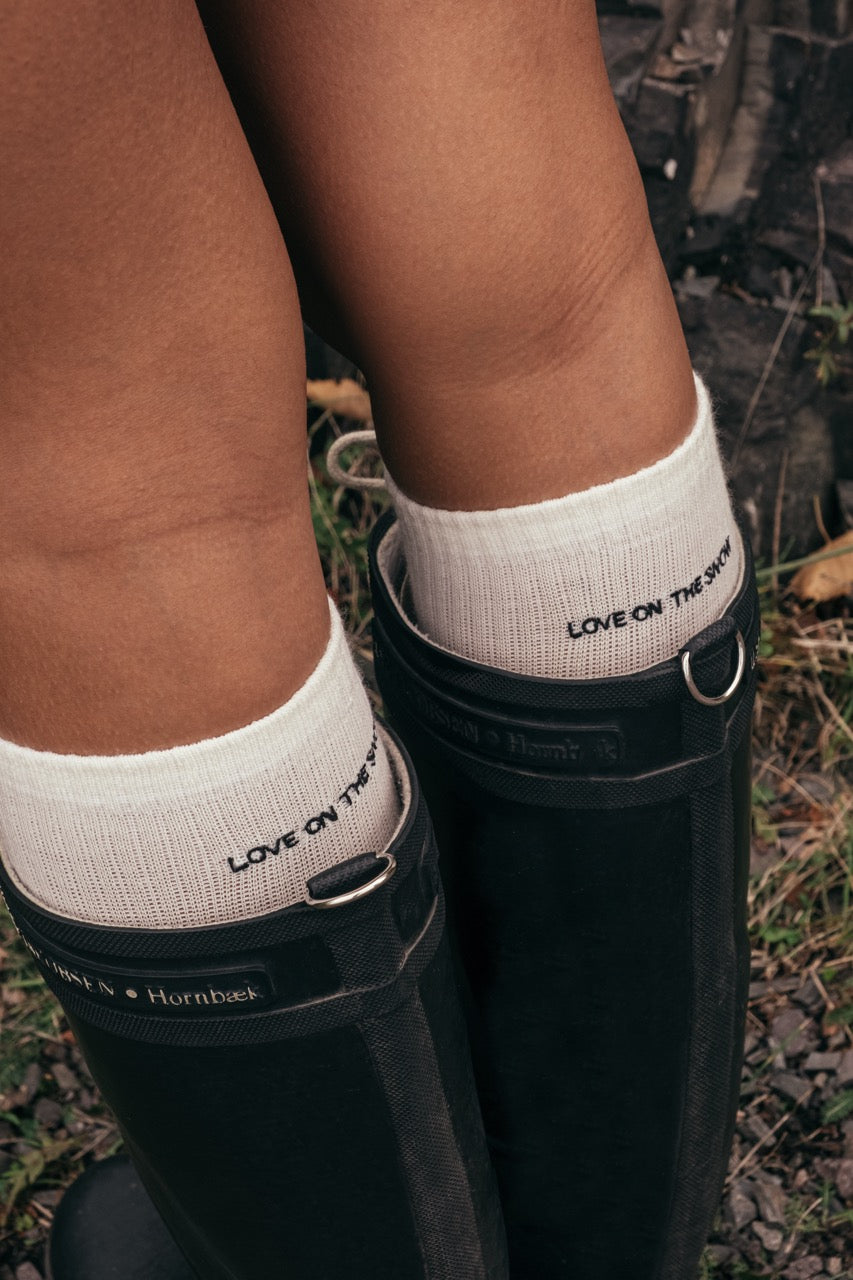 Skarpetki z wełny merino w beżu, z haftowanym logo LOVE ON THE SNOW, na tle wody i krajobrazu.