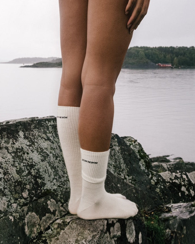 Skarpetki z wełny merino w beżu, z haftowanym logo LOVE ON THE SNOW, na tle wody i krajobrazu.