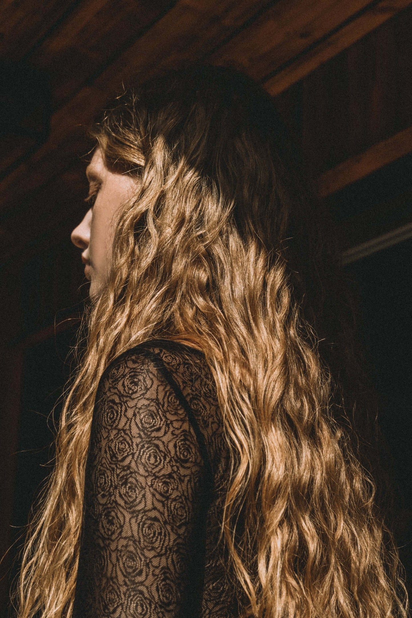 Model with wavy hair wearing a black lace long-sleeve dress with floral patterns, captured in a warm, rustic setting.