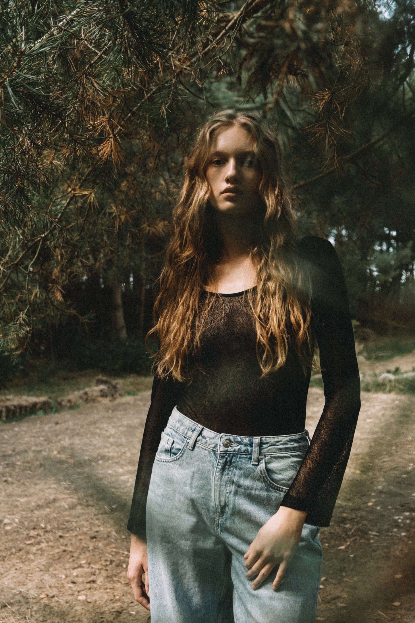 Model wearing a black lace bodysuit with long sleeves, paired with light blue jeans in a forest setting.