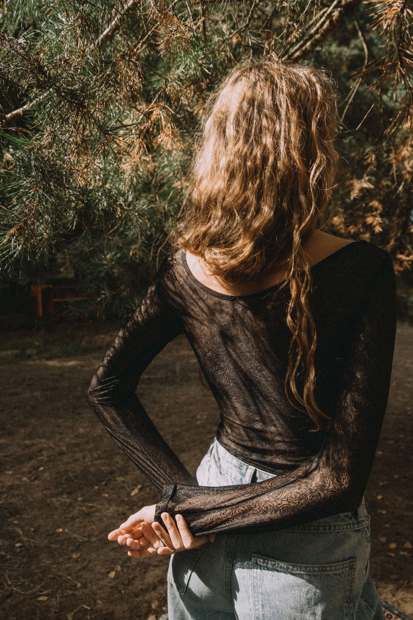 Model wearing a sheer black long-sleeve top standing outdoors, showcasing a casual fashion style.