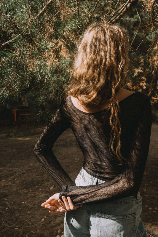Model wearing a sheer black long-sleeve top standing outdoors, showcasing a casual fashion style.