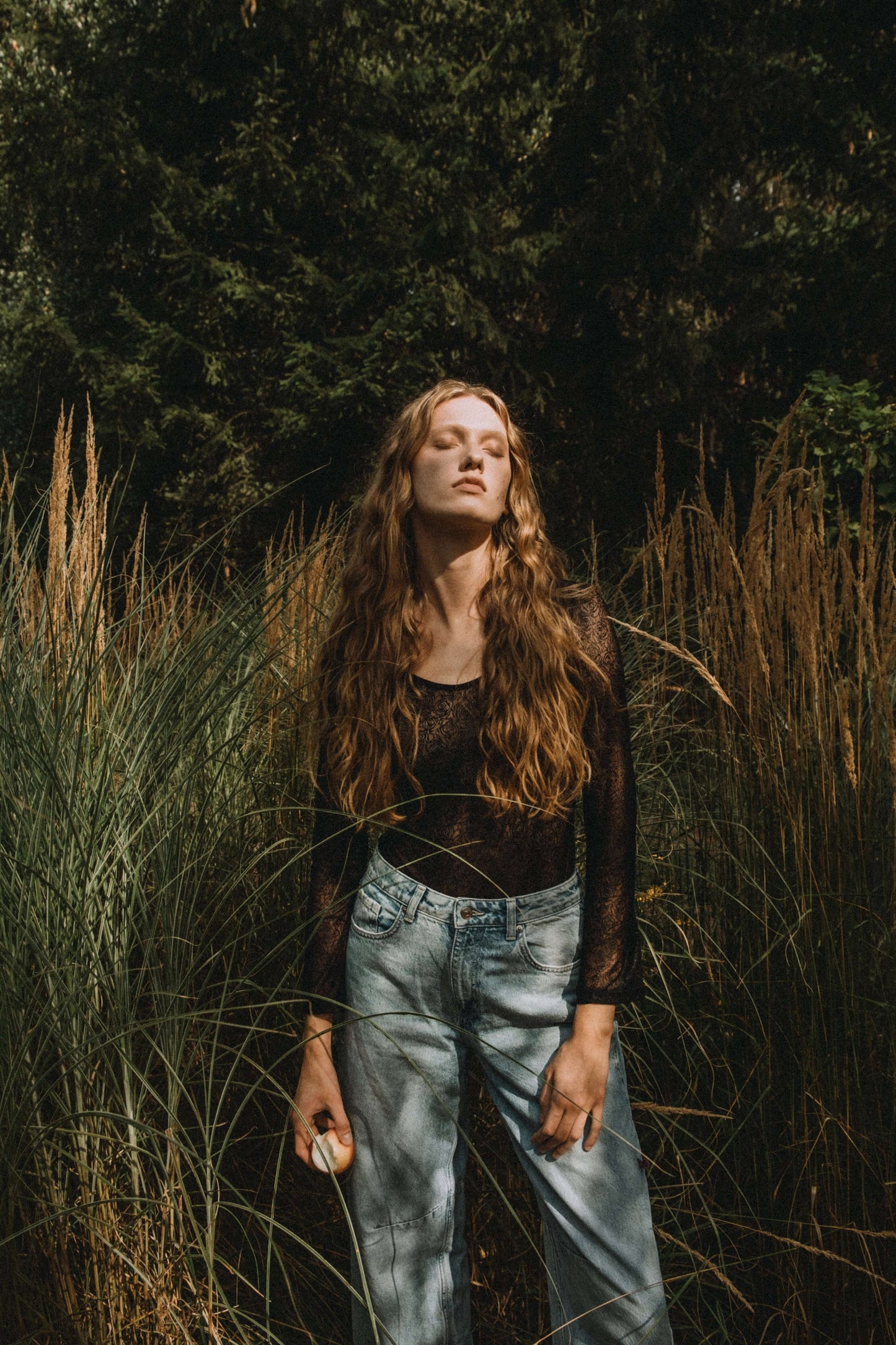 Model in a black lace body top and light jeans standing amidst tall grass in a nature setting.