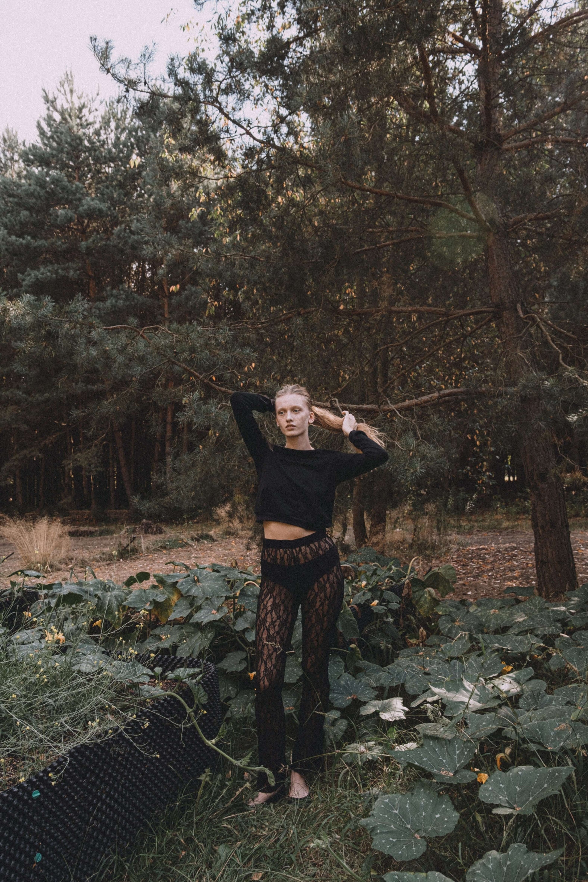 Model in a black crop top and patterned pants poses outdoors among greenery.