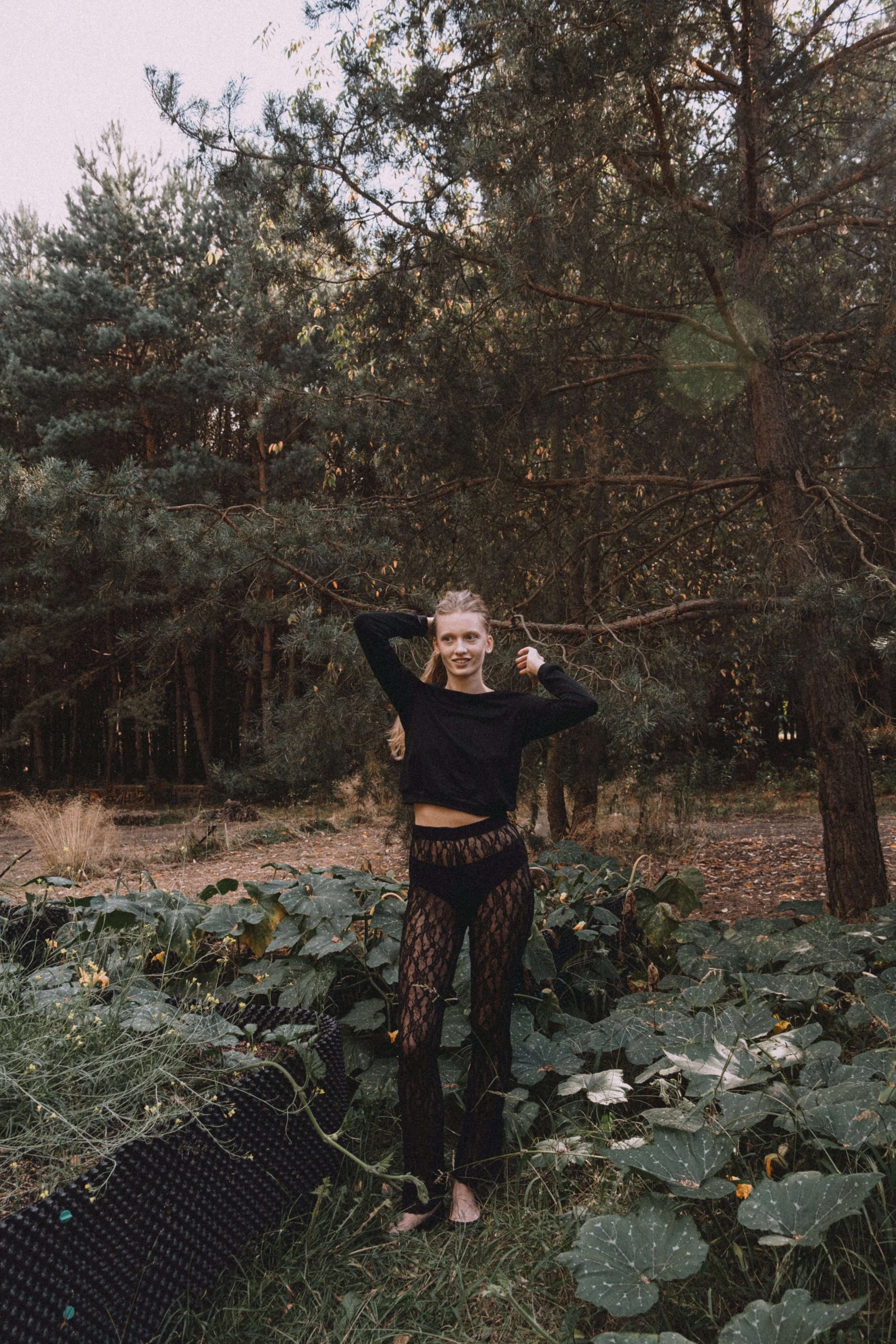 Model wearing a black long-sleeve top and patterned pants, posing outdoors among greenery.