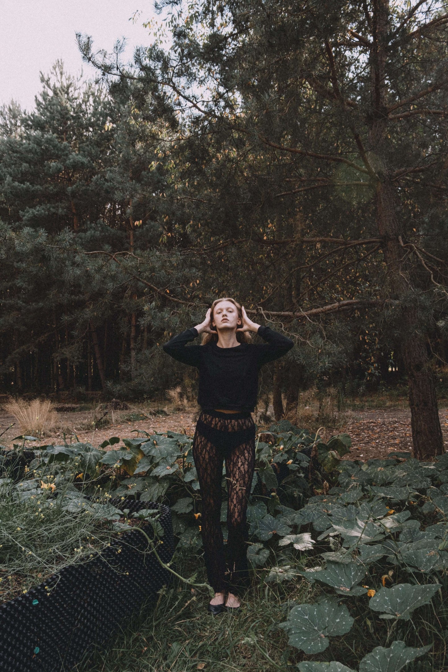 Woman in a black sweater and lace pants posing in a forest surrounded by greenery.