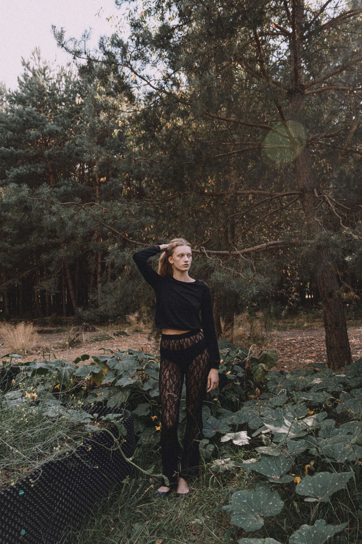 Model in a black long-sleeve top and patterned pants surrounded by greenery in a forest.