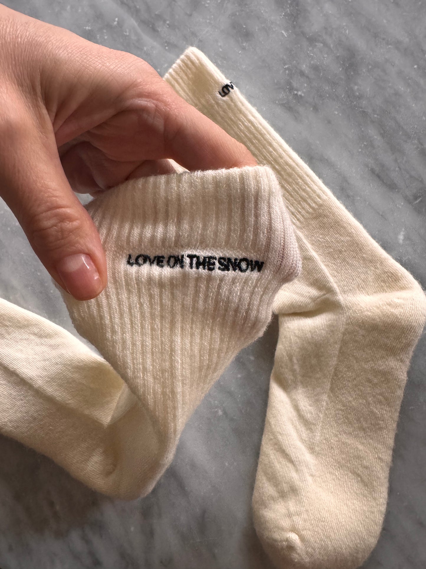 Skarpetki z wełny merino w beżu, z haftowanym logo LOVE ON THE SNOW, na tle wody i krajobrazu.