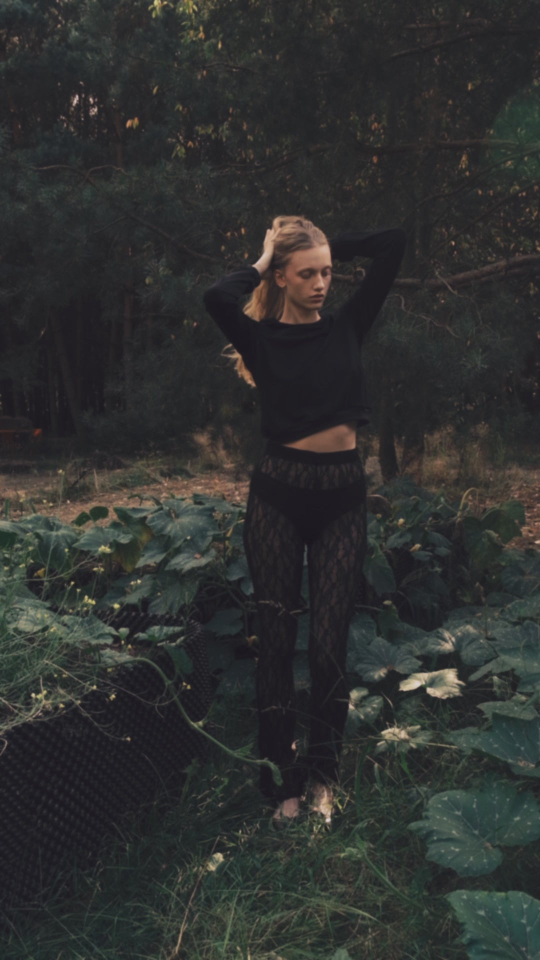Woman in a black cropped sweater and patterned leggings stands in a lush green setting outdoors.