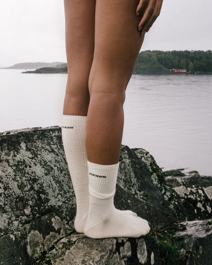 Skarpetki z wełny merino w beżu, z haftowanym logo LOVE ON THE SNOW, na tle wody i krajobrazu.