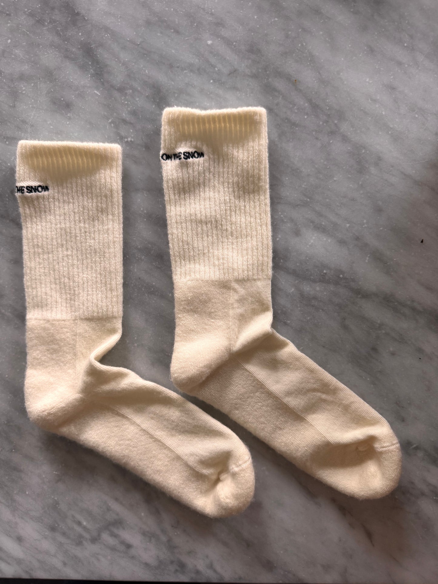 Skarpetki z wełny merino w beżu, z haftowanym logo LOVE ON THE SNOW, na tle wody i krajobrazu.