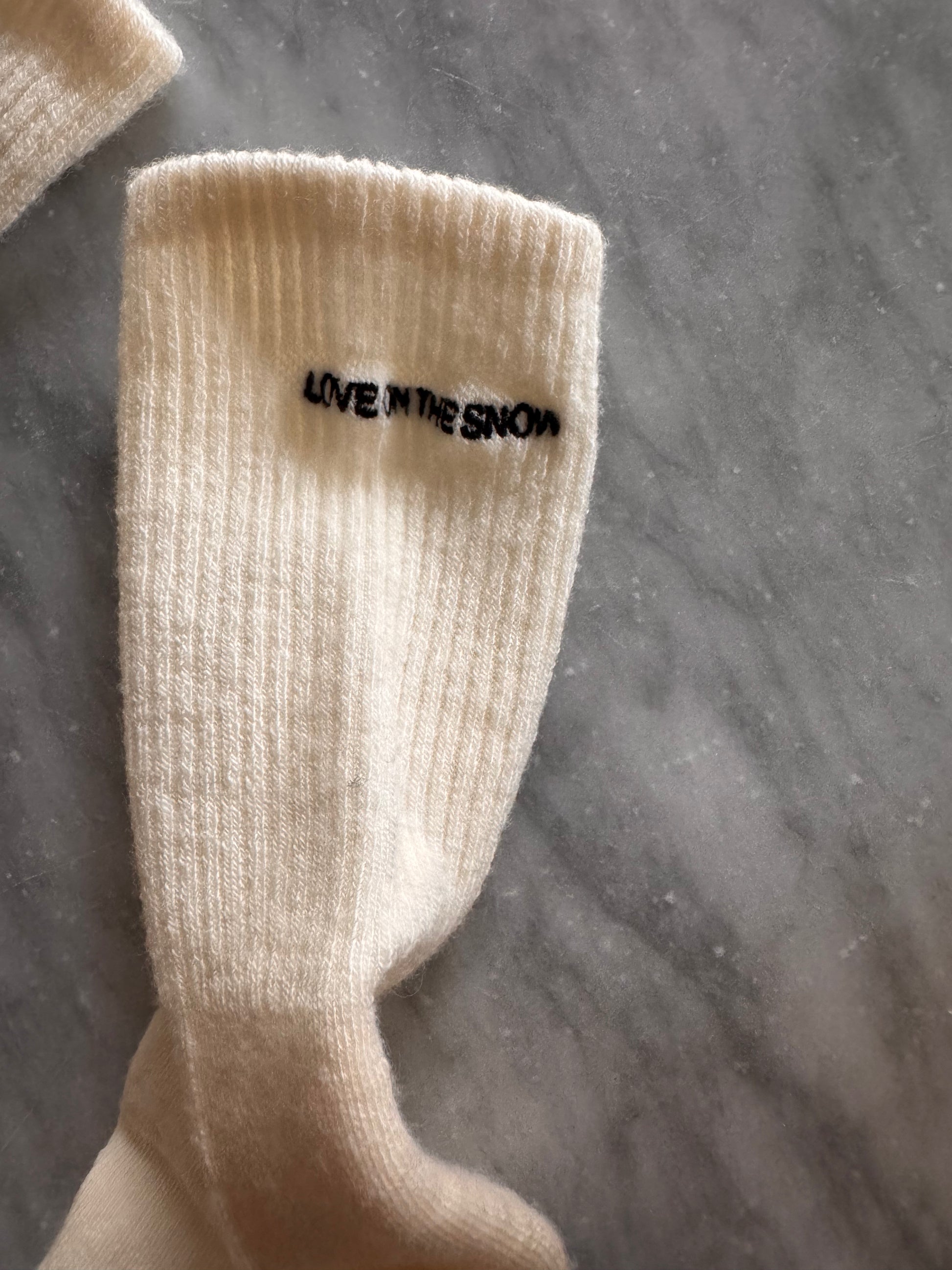 Skarpetki z wełny merino w beżu, z haftowanym logo LOVE ON THE SNOW, na tle wody i krajobrazu.