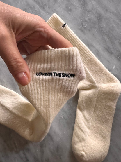 Skarpetki z wełny merino w beżu, z haftowanym logo LOVE ON THE SNOW, na tle wody i krajobrazu.